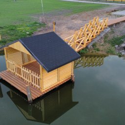 Tartak Zągoty tarasy , patio altany - Drewniana altana z czarnym dachem, zbudowana na wodzie, połączona drewnianym pomostem z brzegiem. Widok z góry, spokojna tafla wody odbija konstrukcję.