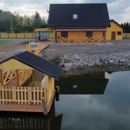 Tartak Zągoty tarasy , patio altany - Drewniana altana na wodzie z balustradą, odbijająca się w tafli stawu. W tle dom z drewna i żółte ogrodzenie. Pochmurne niebo.