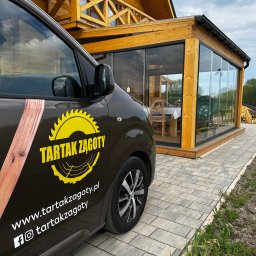 Tartak Zągoty tarasy , patio altany - Auto z logo Tartak Żagoty na tle drewnianej konstrukcji z przeszkloną werandą i balkonem. Widoczny fragment kostki brukowej i fragment trawnika.