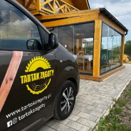 Auto z logo Tartak Żagoty na tle drewnianej konstrukcji z przeszkloną werandą i balkonem. Widoczny fragment kostki brukowej i fragment trawnika.