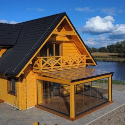 Tartak Zągoty tarasy , patio altany - Żółty dom z drewna z przeszklonym tarasem i balkonem, czarnym dachem, położony nad jeziorem w słoneczny dzień. Widok z góry ukazuje detale konstrukcji i otoczenie.