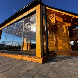 Nowoczesna, drewniana konstrukcja z przeszkleniami i pergolą w tle, widziana z poziomu brukowanej nawierzchni w słoneczny dzień. Detal architektoniczny.