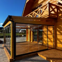 Drewniany dom z przeszklonym tarasem i balkonem, widok z zewnątrz w słoneczny dzień. Czarna rynna, drewniane wykończenie, kostka brukowa na pierwszym planie.