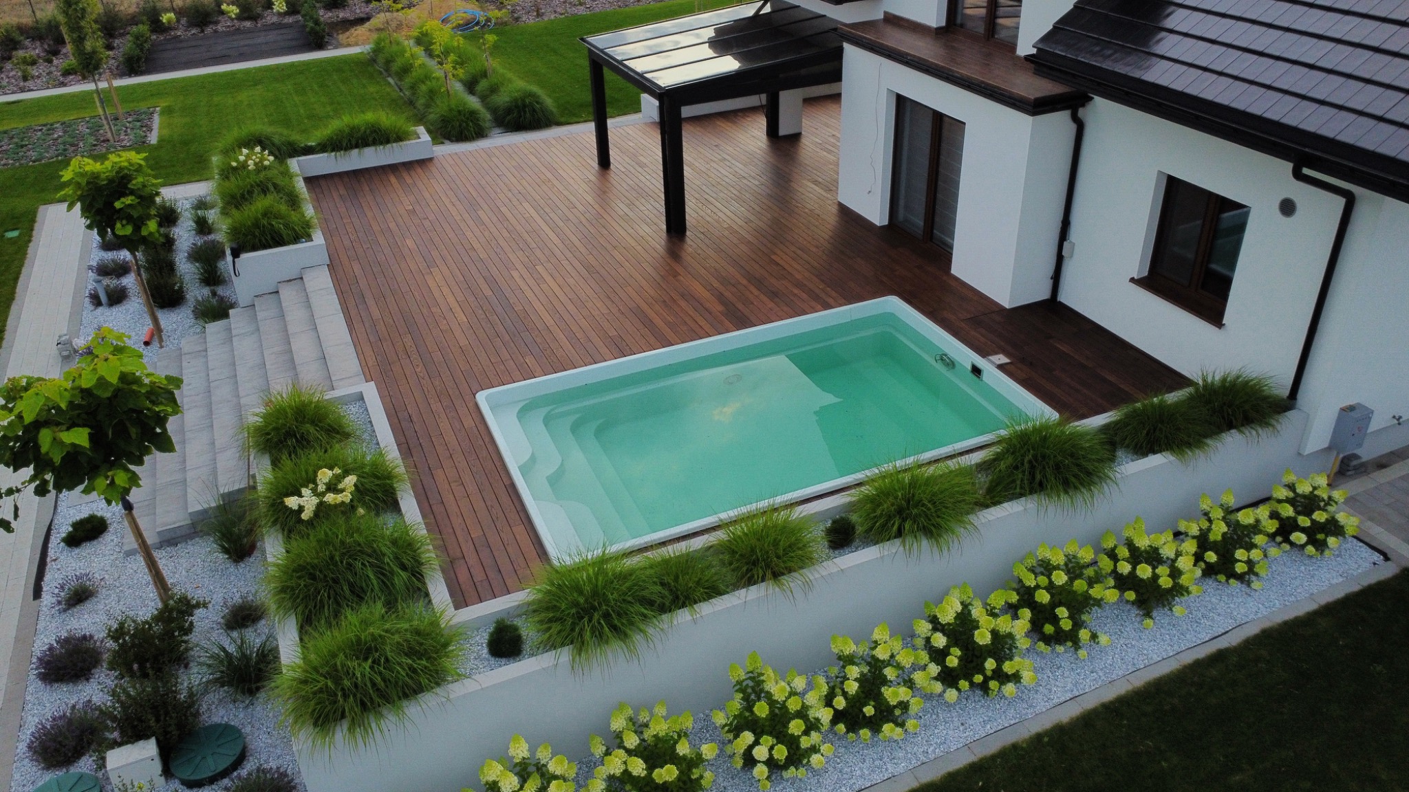 Nowoczesny taras z drewna egzotycznego, zintegrowany z basenem i otoczony zielenią. Widok z góry ukazuje harmonijną kompozycję przestrzeni wypoczynkowej.