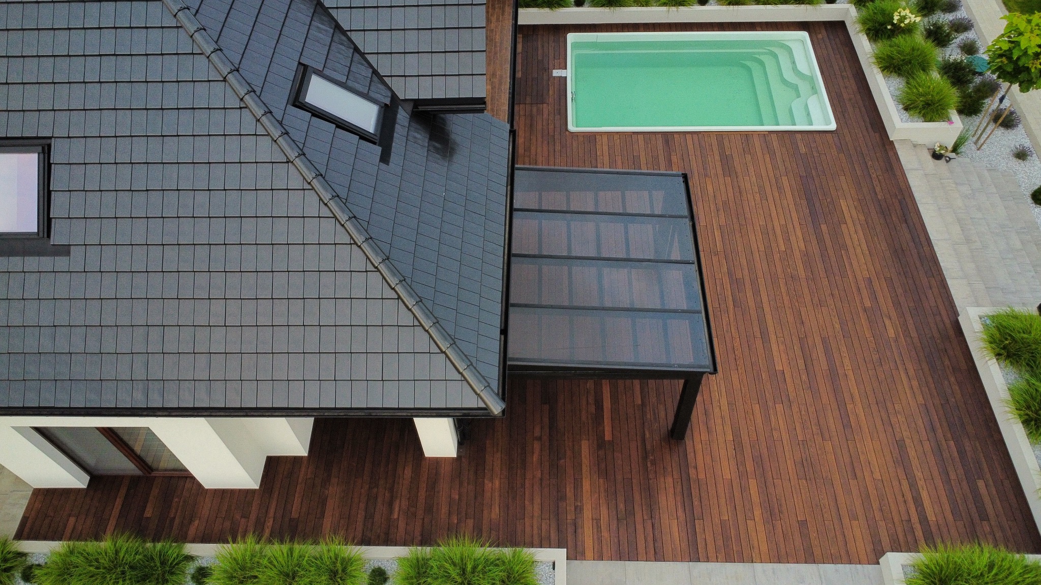 Widok z góry na taras z drewna egzotycznego z basenem i zadaszeniem, przylegający do domu z ciemną dachówką. Uwagę przykuwa precyzyjny montaż desek i estetyczne wykończenie.