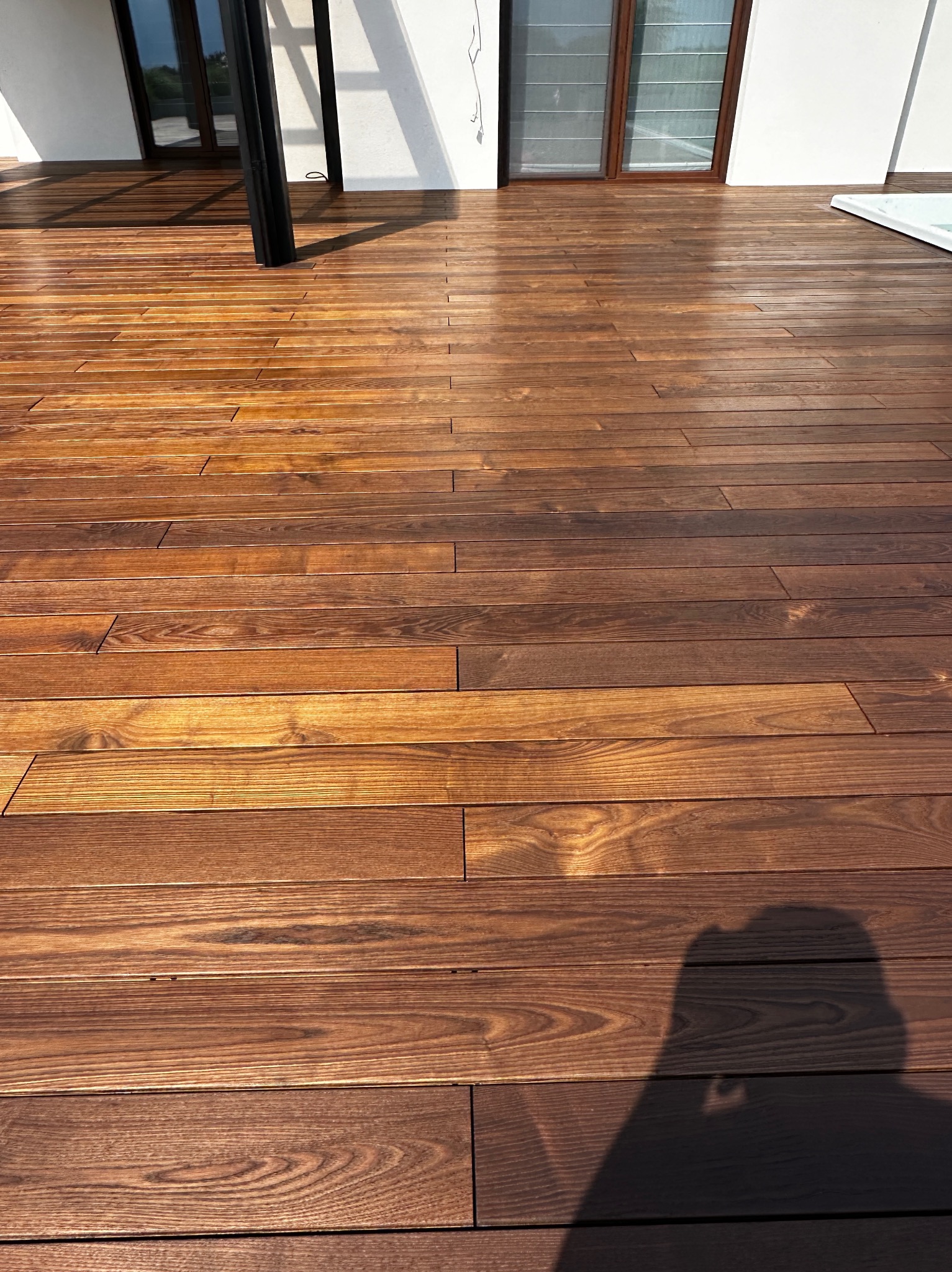 Błyszczący, brązowy taras z drewna egzotycznego, z widoczną strukturą słojów, oświetlony naturalnym światłem, z fragmentem białej ściany i czarnego słupa w tle.