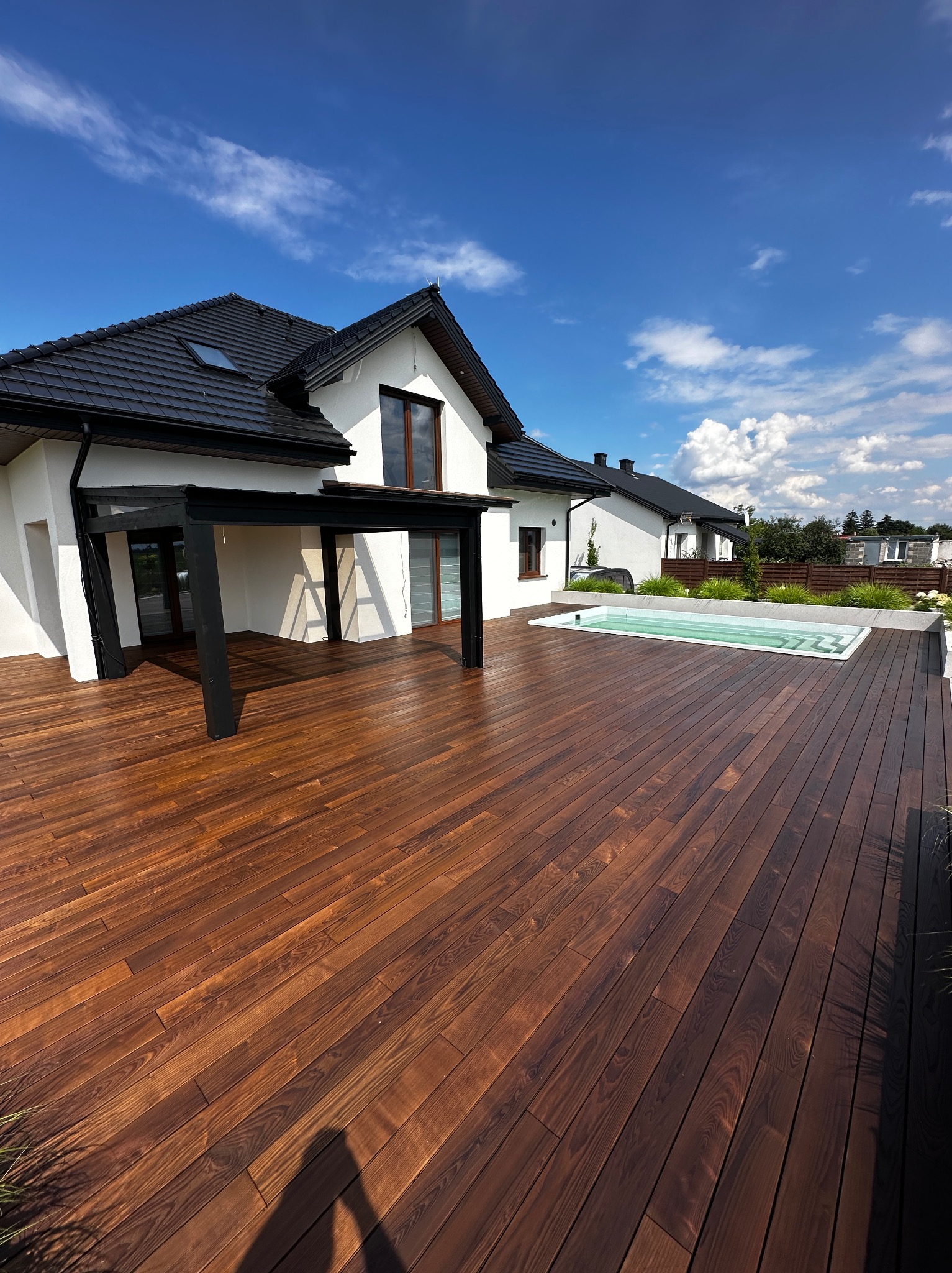 Nowoczesny taras z ciemnego drewna otaczający basen przy białym domu z czarnym dachem, błękitne niebo z chmurami w tle. Ujęcie z niskiej perspektywy.