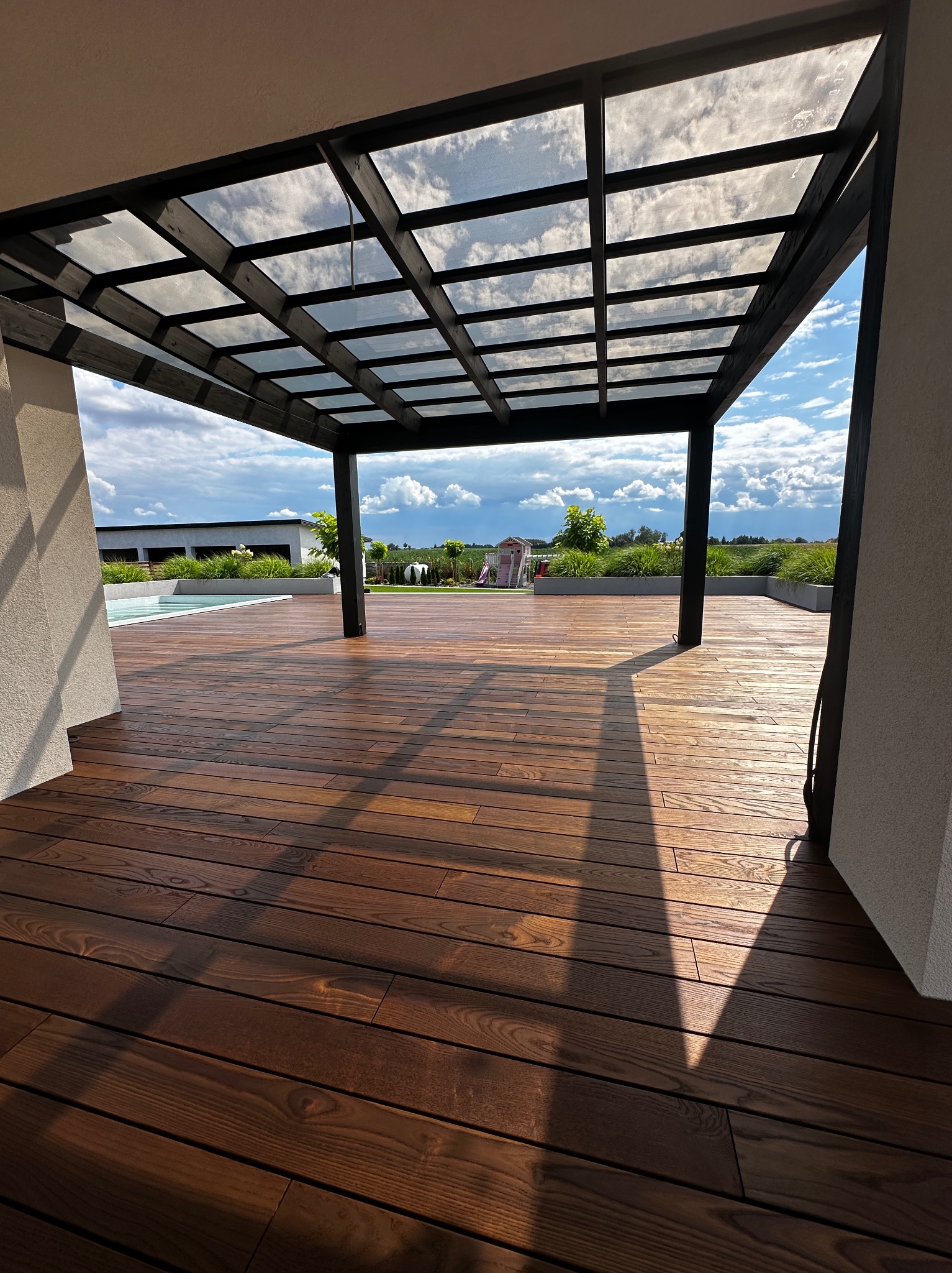 Nowoczesny taras z ciemnego drewna pod pergolą z przezroczystym zadaszeniem, widok na ogród i basen w słoneczny dzień.