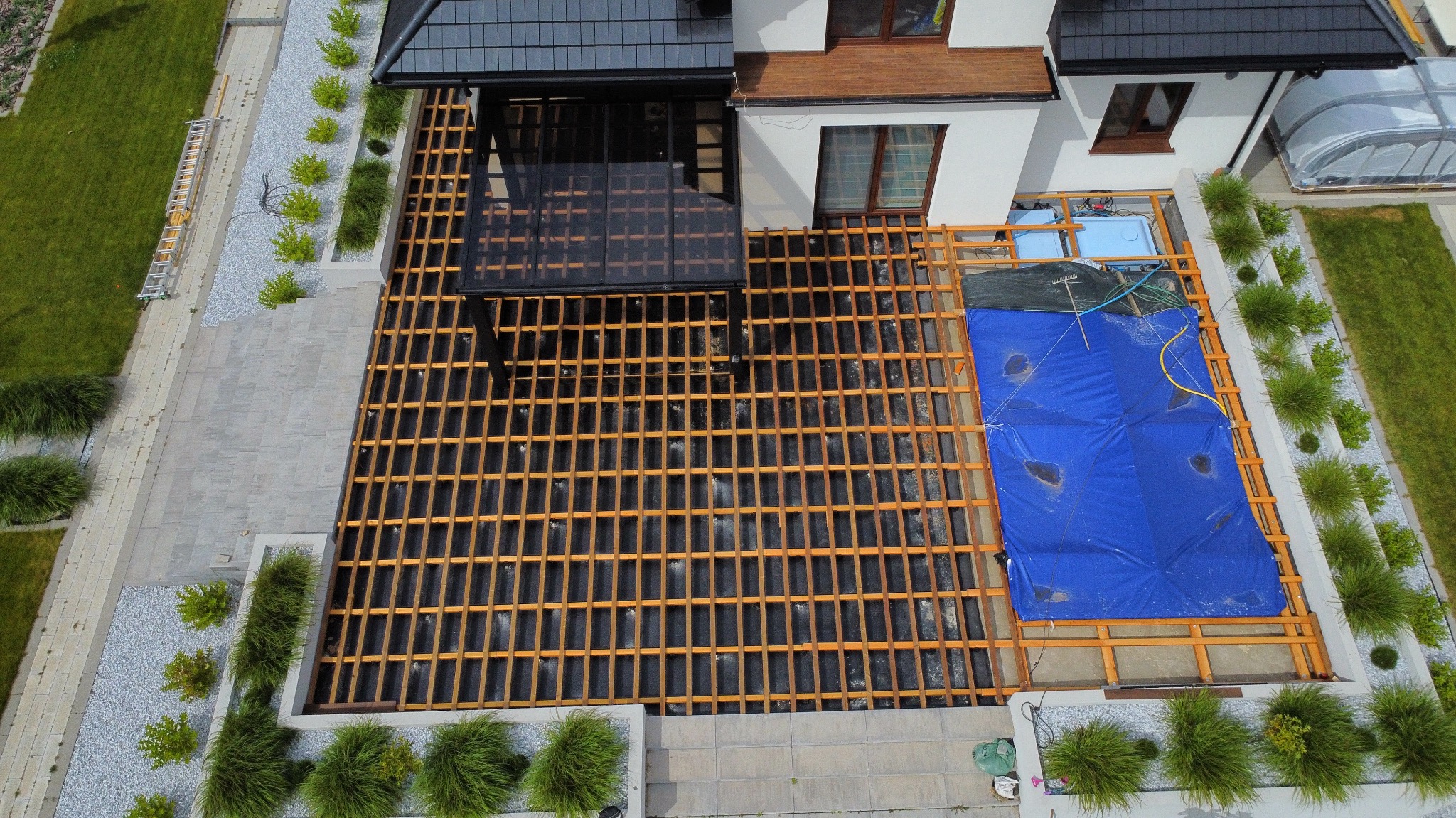 Widok z góry na konstrukcję tarasu w trakcie budowy, z elementami przykrytymi plandeką. Obok domu widoczny ogród z dekoracyjnymi kamieniami i roślinnością.