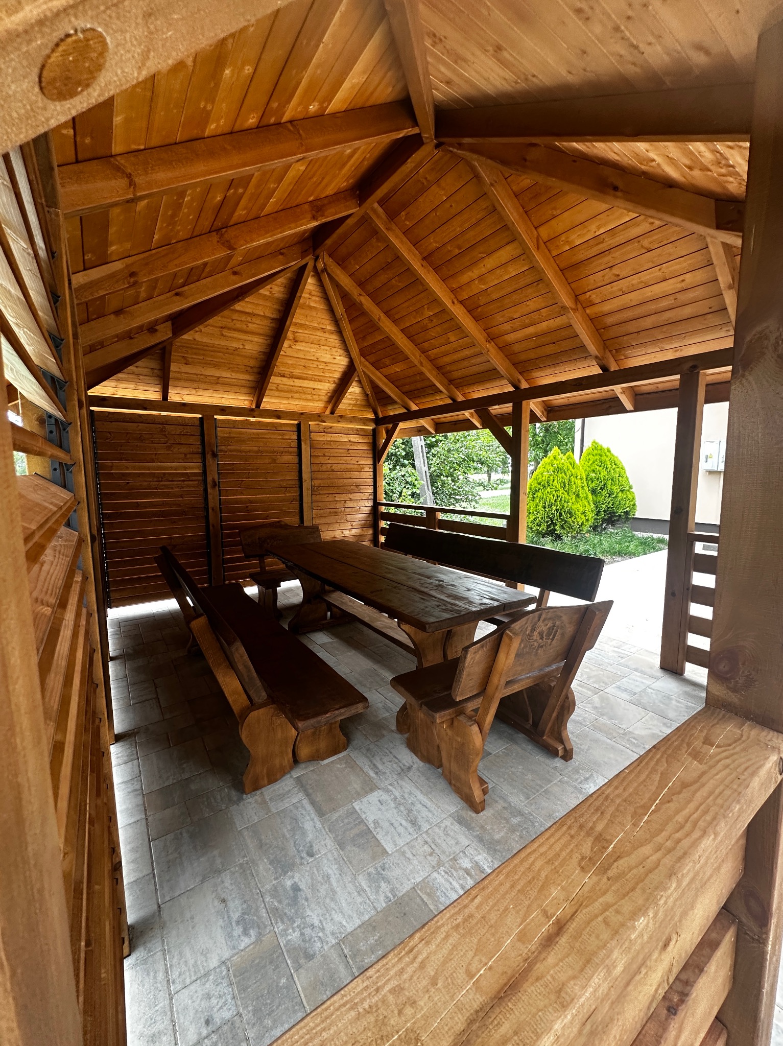 Drewniana altana z masywnym stołem i ławami, widok z zewnątrz. Konstrukcja z jasnego drewna, zadaszenie skośne, częściowo zabudowane ściany, podłoga wyłożona płytkami.