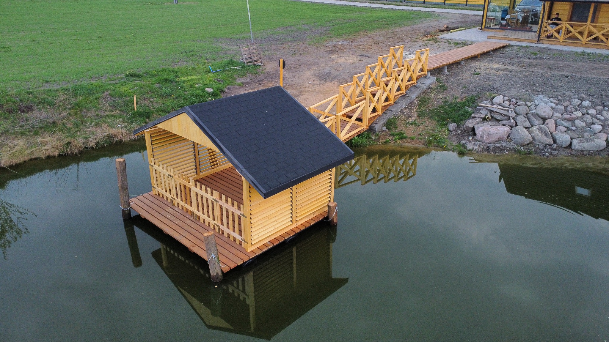 Drewniana altana z czarnym dachem, zbudowana na wodzie, połączona drewnianym pomostem z brzegiem. Widok z góry, spokojna tafla wody odbija konstrukcję.