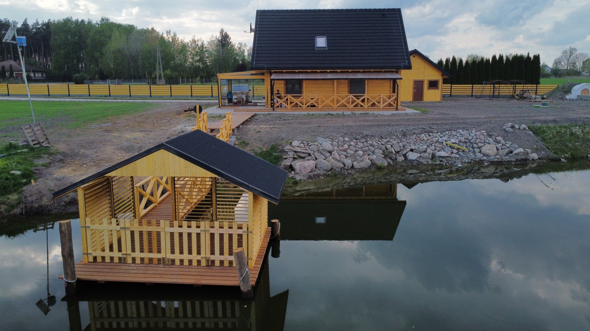 Drewniana altana na wodzie z balustradą, odbijająca się w tafli stawu. W tle dom z drewna i żółte ogrodzenie. Pochmurne niebo.