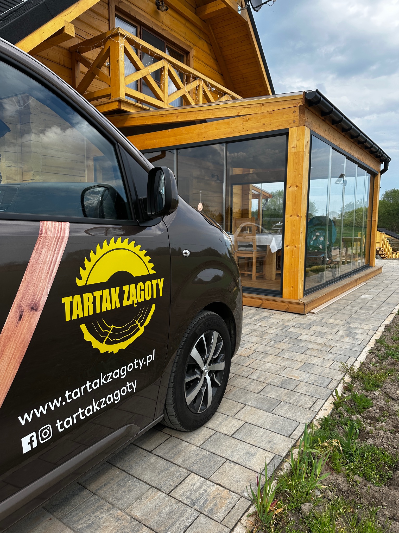 Auto z logo Tartak Żagoty na tle drewnianej konstrukcji z przeszkloną werandą i balkonem. Widoczny fragment kostki brukowej i fragment trawnika.