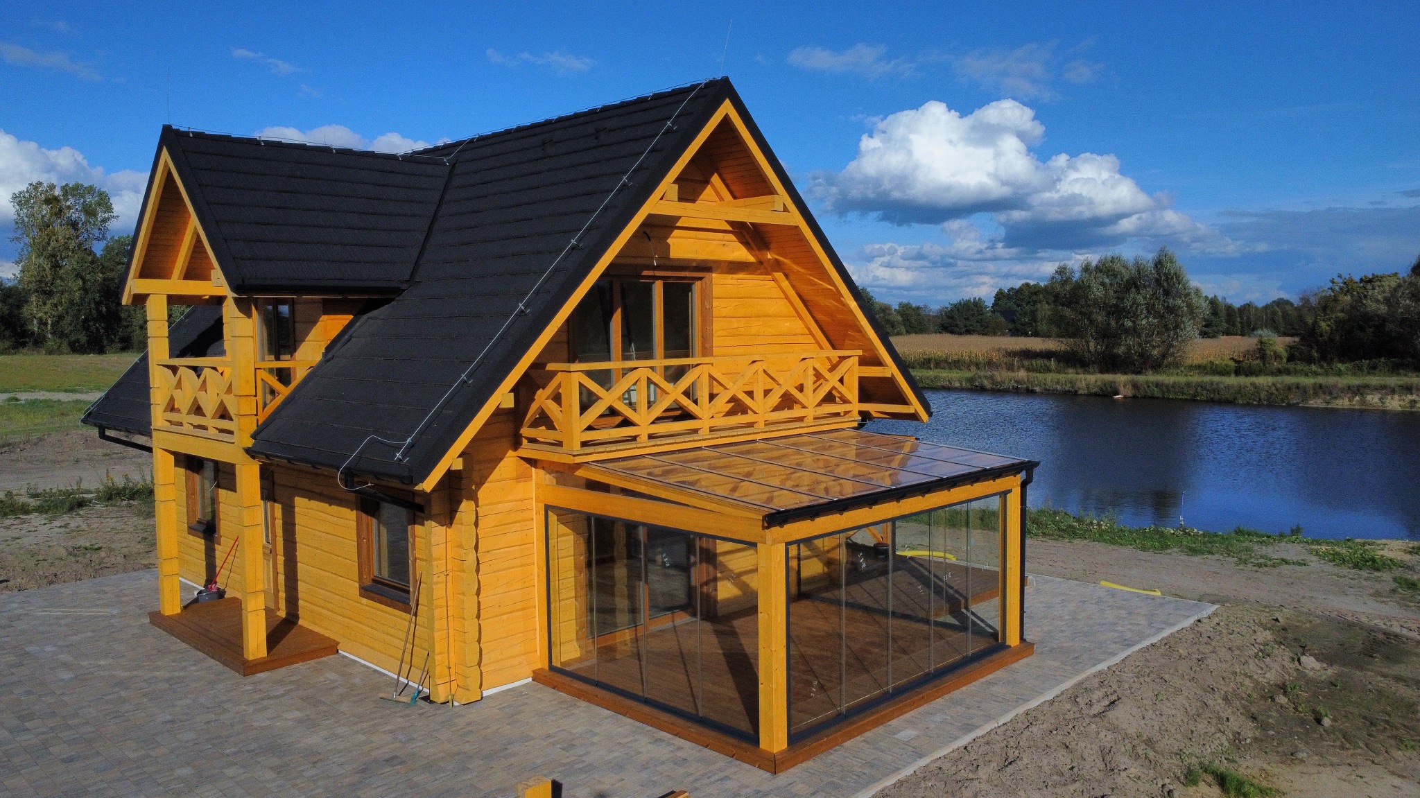 Żółty dom z drewna z przeszklonym tarasem i balkonem, czarnym dachem, położony nad jeziorem w słoneczny dzień. Widok z góry ukazuje detale konstrukcji i otoczenie.