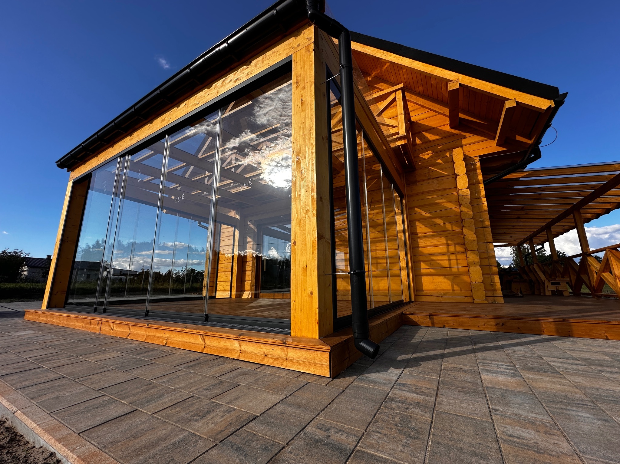 Nowoczesna, drewniana konstrukcja z przeszkleniami i pergolą w tle, widziana z poziomu brukowanej nawierzchni w słoneczny dzień. Detal architektoniczny.