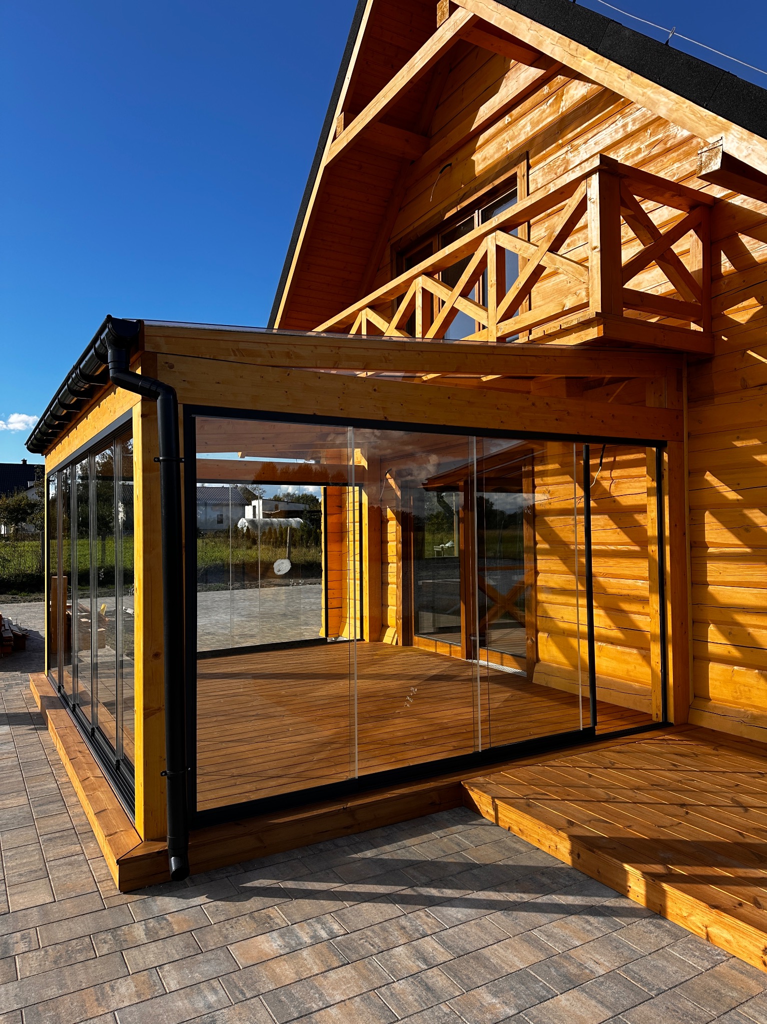 Drewniany dom z przeszklonym tarasem i balkonem, widok z zewnątrz w słoneczny dzień. Czarna rynna, drewniane wykończenie, kostka brukowa na pierwszym planie.