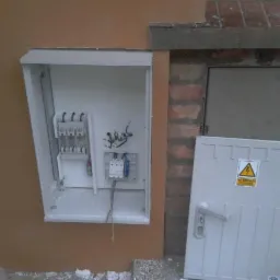 Otwarta, biała skrzynka elektryczna zamontowana na pomarańczowej ścianie budynku, z widocznymi elementami instalacji wewnątrz i uchyloną obok pokrywą z naklejką ostrzegawczą przed wysokim napięciem.