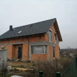 Dwukondygnacyjny budynek w stanie surowym otwartym, z czarną dachówką, oknami zabezpieczonymi folią, ceglanymi ścianami i widocznymi elementami konstrukcyjnymi, otoczony zielonym ogrodzeniem...