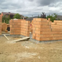 Wznoszenie ścian z czerwonej cegły na fundamencie. Widoczne fragmenty murów, palety z cegłami i drewniane deski. W tle domy i pochmurne niebo.