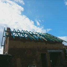 Wznoszenie więźby dachowej z impregnowanego drewna na murowanym budynku w słoneczny dzień. Widoczne elementy konstrukcyjne i niebieskie niebo z chmurami.