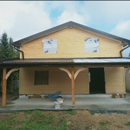 Surowy drewniany dom z czarnym dachem i rynnami w trakcie budowy, z widoczną konstrukcją zadaszenia tarasu i materiałami budowlanymi na betonowej wylewce.