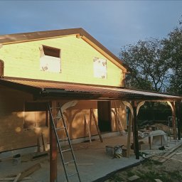 Drewniany dom w trakcie budowy z widoczną konstrukcją dachu i zadaszeniem tarasu. Na pierwszym planie drabina i materiały budowlane.