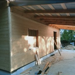 Drewniana konstrukcja z widoczną więźbą dachową i deskami elewacyjnymi w trakcie montażu. Widoczne narzędzia i materiały budowlane. Częściowo zadaszona konstrukcja.