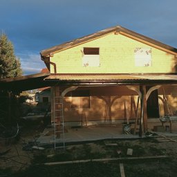 Usługi Ciesielskie, Dekarskie,Remontowo-Budowlane i Tartaczne TOMASZ BEBŁOT - Dom w trakcie budowy z widoczną drewnianą konstrukcją dachu i ścian. Na pierwszym planie zadaszenie tarasu. W tle ciemne chmury.