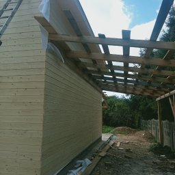 Usługi Ciesielskie, Dekarskie,Remontowo-Budowlane i Tartaczne TOMASZ BEBŁOT - Budowa drewnianej konstrukcji z widoczną więźbą dachową i deskowaniem ścian. Na ziemi leżą deski i inne materiały budowlane. W tle zieleń i błękitne niebo.