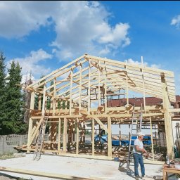 Usługi Ciesielskie, Dekarskie,Remontowo-Budowlane i Tartaczne TOMASZ BEBŁOT - Wznoszenie drewnianej konstrukcji domu jednorodzinnego na tle błękitnego nieba z chmurami. Widoczne elementy więźby dachowej i szkieletu budynku. Prace budowlane w toku.