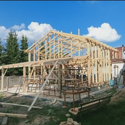 Usługi Ciesielskie, Dekarskie,Remontowo-Budowlane i Tartaczne TOMASZ BEBŁOT - Wznoszenie drewnianej konstrukcji domu jednorodzinnego w słoneczny dzień. Widoczna więźba dachowa w trakcie montażu, elementy rusztowania i pracownik przy pracy.