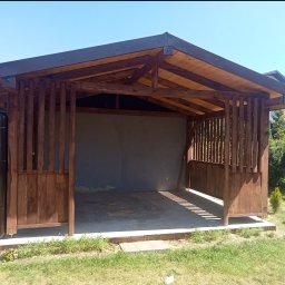 Usługi Ciesielskie, Dekarskie,Remontowo-Budowlane i Tartaczne TOMASZ BEBŁOT - Drewniana wiata ogrodowa z ciemnym dachem, widok z przodu, z elementami dekoracyjnymi z pionowych listew. W tle zieleń ogrodu i budynek gospodarczy.