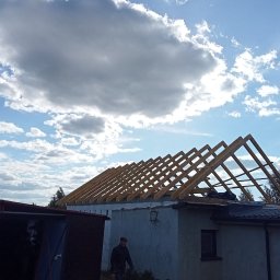 Usługi Ciesielskie, Dekarskie,Remontowo-Budowlane i Tartaczne TOMASZ BEBŁOT - Wznoszenie drewnianej więźby dachowej na murowanym budynku w słoneczny dzień. Widoczne elementy konstrukcyjne i osoba w oddali. Prace dekarskie w toku.