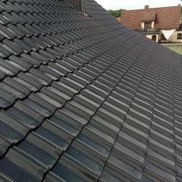Usługi Ciesielskie, Dekarskie,Remontowo-Budowlane i Tartaczne TOMASZ BEBŁOT - Błyszczący, czarny dach z dachówki ceramicznej w ukośnym ujęciu. W tle widoczny budynek mieszkalny z brązowym dachem i fragment zieleni.