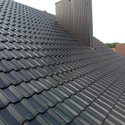 Usługi Ciesielskie, Dekarskie,Remontowo-Budowlane i Tartaczne TOMASZ BEBŁOT - Błyszczący, grafitowy dach z blachodachówki trapezowej i komin z blachy na tle jasnego nieba. Widok z góry, ukośny kadr.