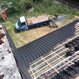 Usługi Ciesielskie, Dekarskie,Remontowo-Budowlane i Tartaczne TOMASZ BEBŁOT - Montaż czarnej dachówki na drewnianej konstrukcji dachu. Widoczny pracownik przy pracy, drabina i auto dostawcze. Ujęcie z góry, w tle zieleń.