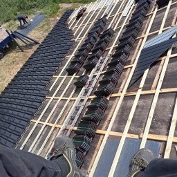 Usługi Ciesielskie, Dekarskie,Remontowo-Budowlane i Tartaczne TOMASZ BEBŁOT - Montaż czarnej dachówki na drewnianej konstrukcji dachu, widok z góry z perspektywy dekarza. Częściowo pokryty dach, drabina i narzędzia w tle.