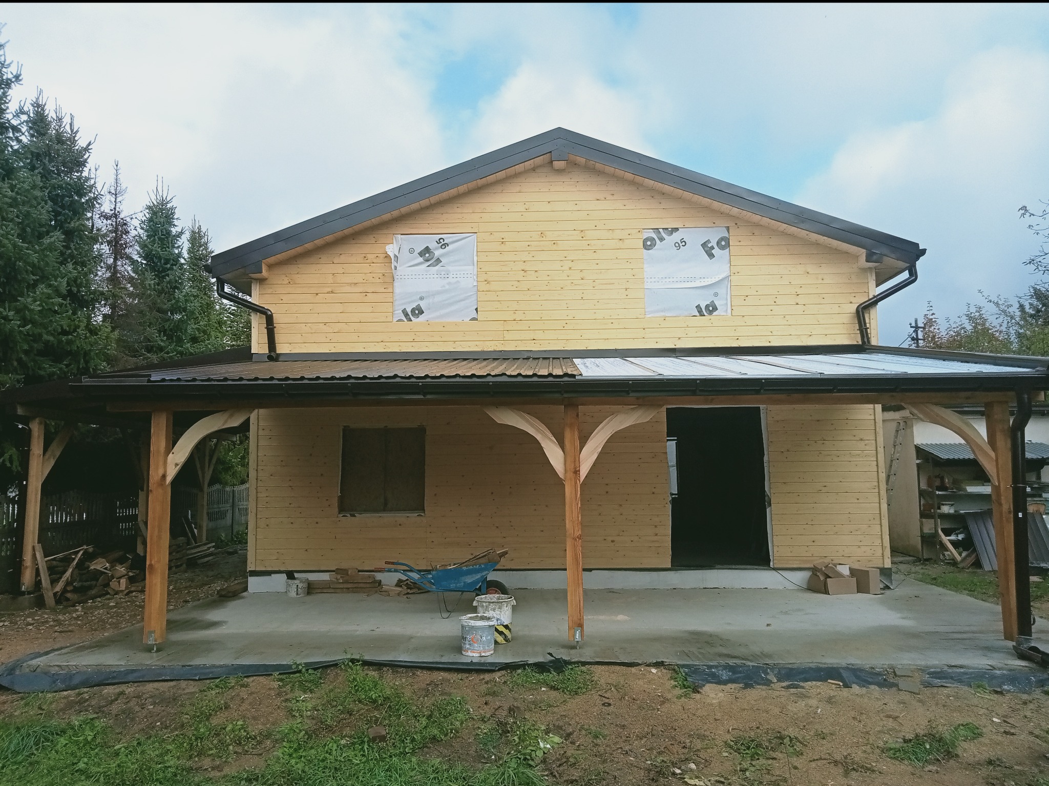 Surowy drewniany dom z czarnym dachem i rynnami w trakcie budowy, z widoczną konstrukcją zadaszenia tarasu i materiałami budowlanymi na betonowej wylewce.