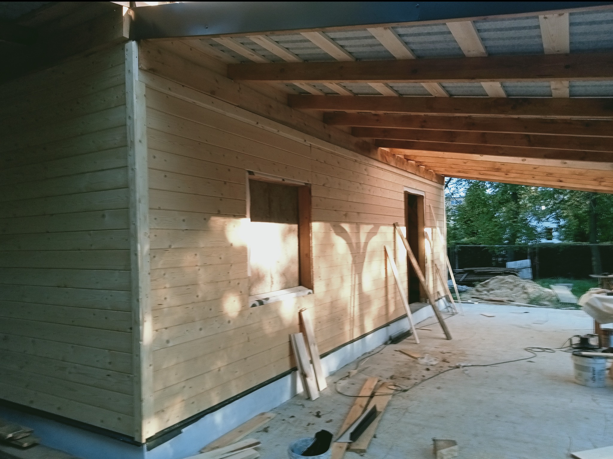 Drewniana konstrukcja z widoczną więźbą dachową i deskami elewacyjnymi w trakcie montażu. Widoczne narzędzia i materiały budowlane. Częściowo zadaszona konstrukcja.