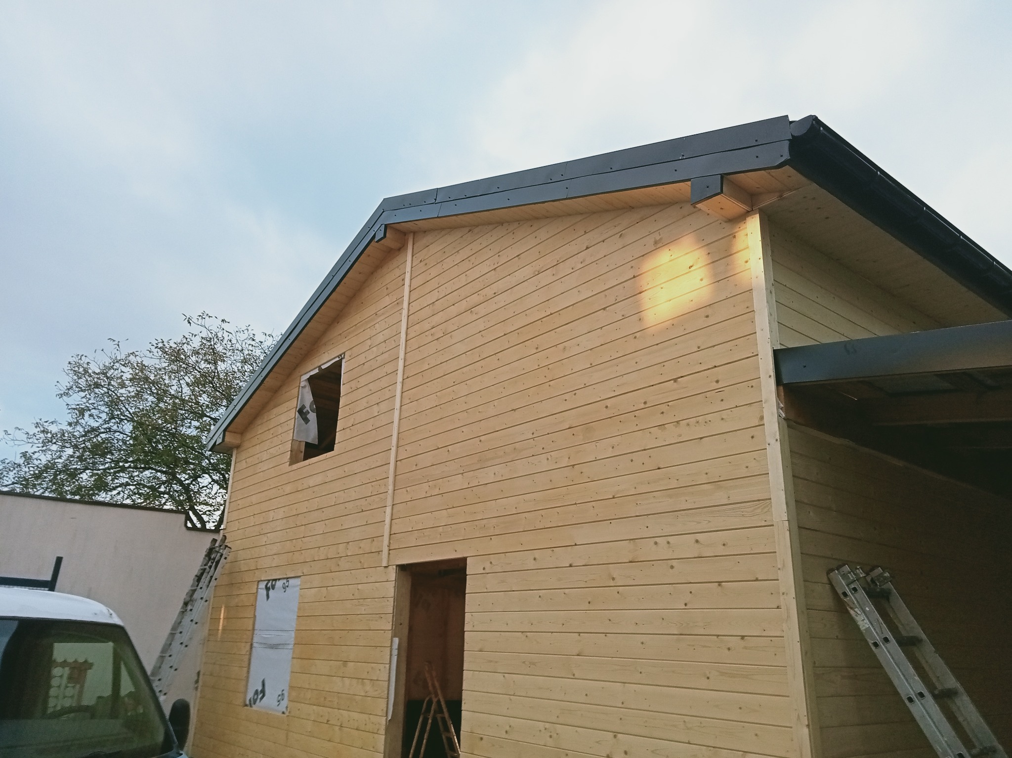 Drewniany dom w trakcie budowy, widoczne deski elewacyjne, okno z folią, drabiny. Ujęcie z dołu, jasne drewno, czarne obróbki dachowe.