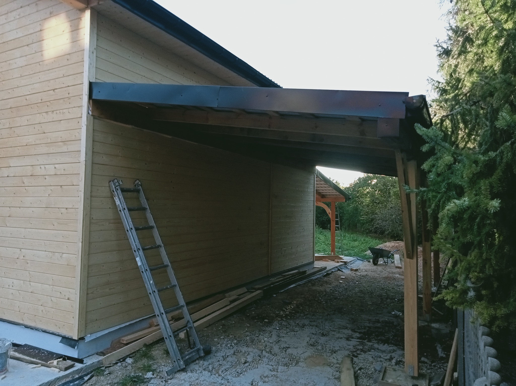 Drewniana konstrukcja z elementami dachu w trakcie budowy, widoczna drabina oparta o ścianę. Ujęcie z dołu, z boku, w tle zieleń.