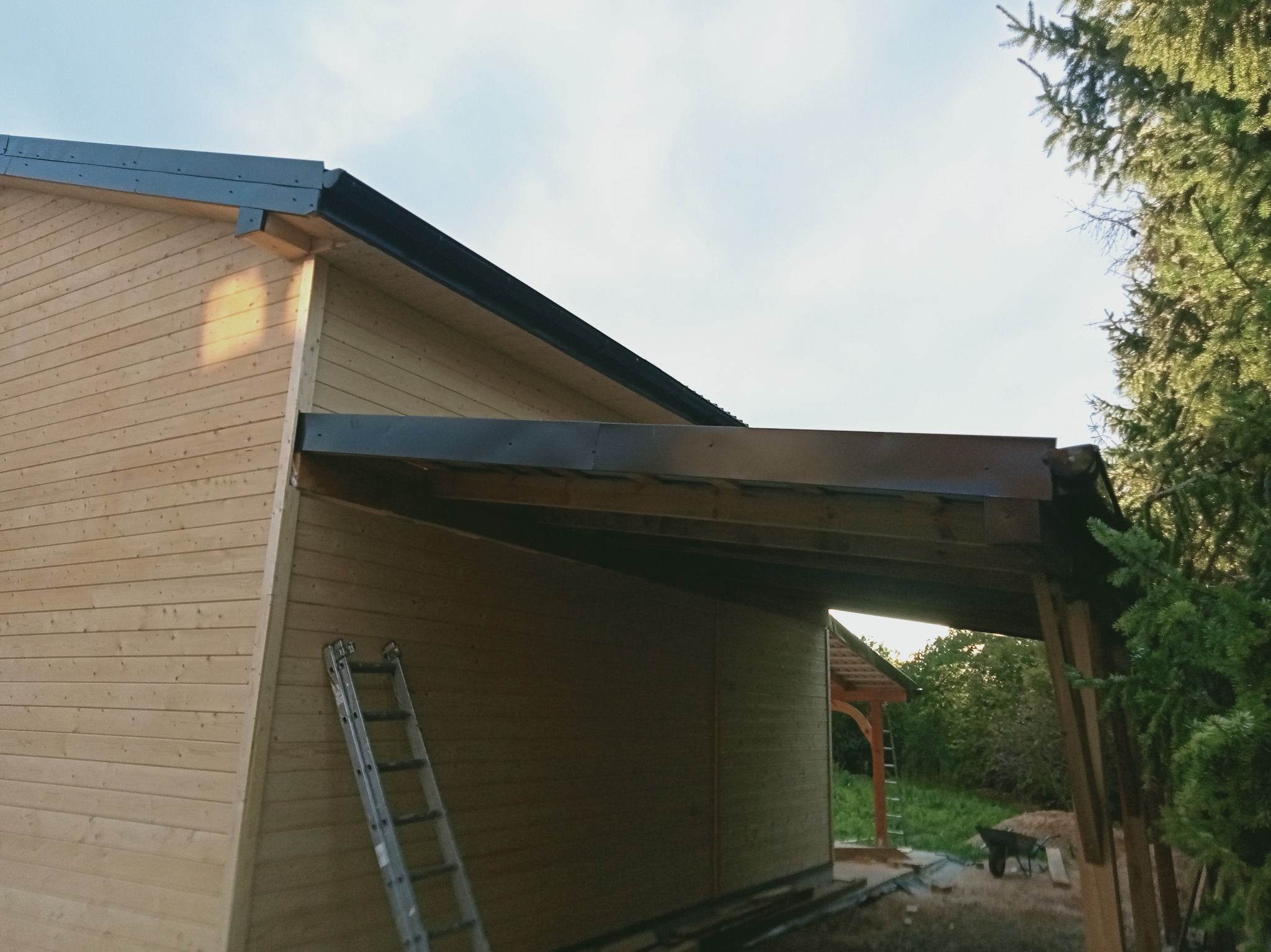 Drewniana konstrukcja dachu w trakcie budowy, widoczna drabina oparta o ścianę, elementy więźby dachowej i fragment otoczenia z zielenią.