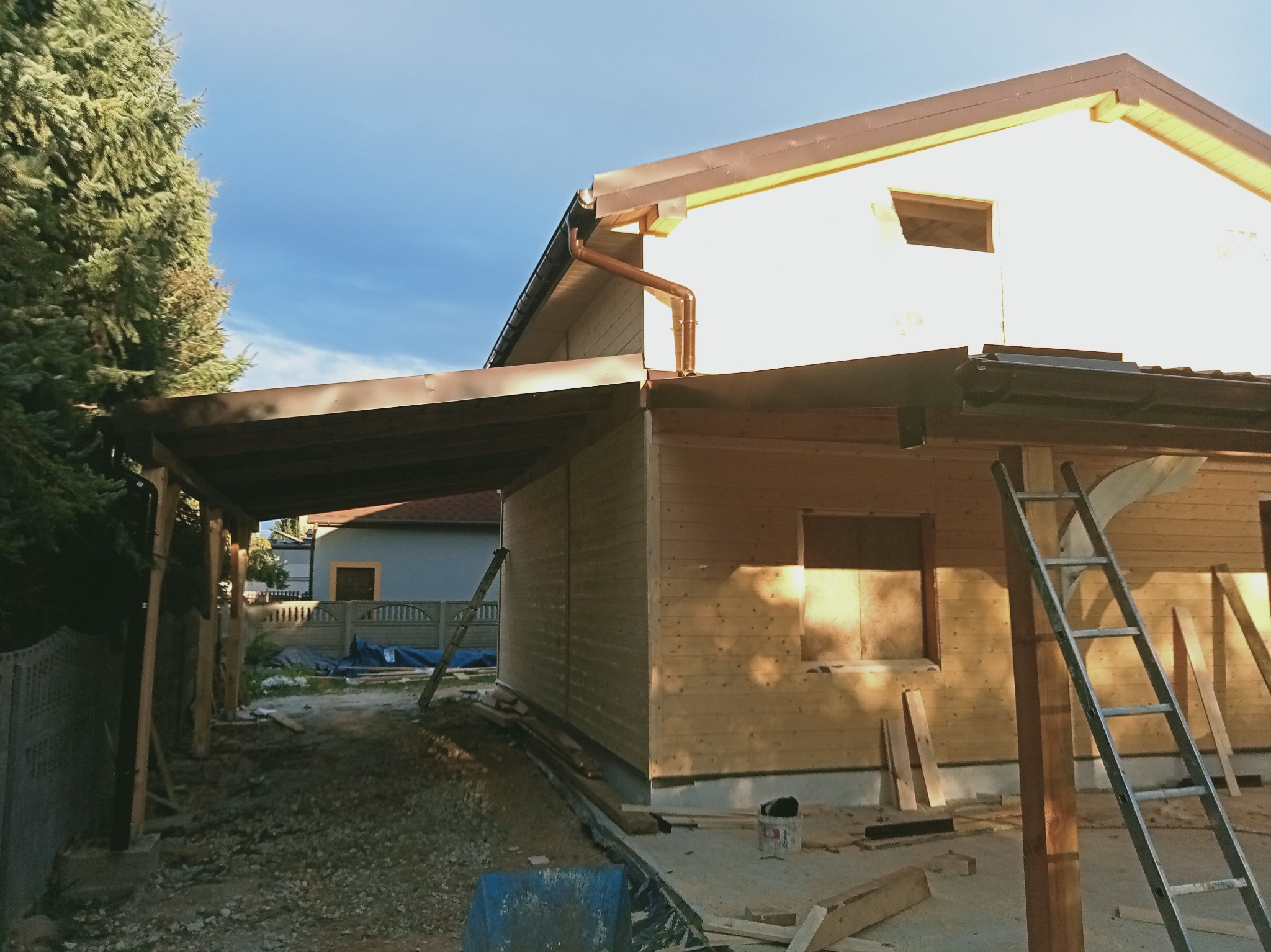 Drewniana konstrukcja z brązowymi rynnami i zadaszeniem, częściowo ukończona, z drabiną opartą o ścianę. Widoczne narzędzia i materiały budowlane.
