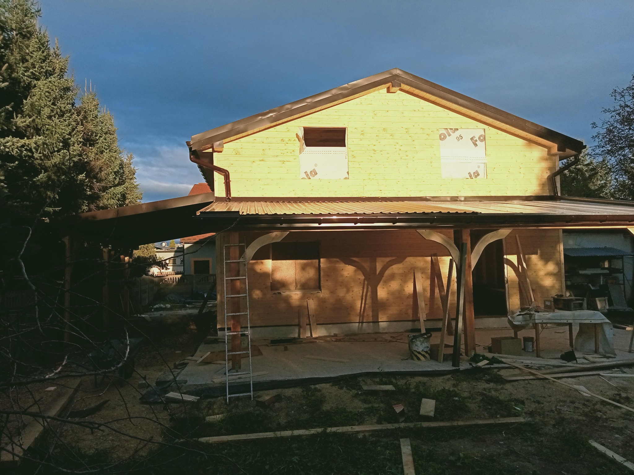 Dom w trakcie budowy z widoczną drewnianą konstrukcją dachu i ścian. Na pierwszym planie zadaszenie tarasu. W tle ciemne chmury.