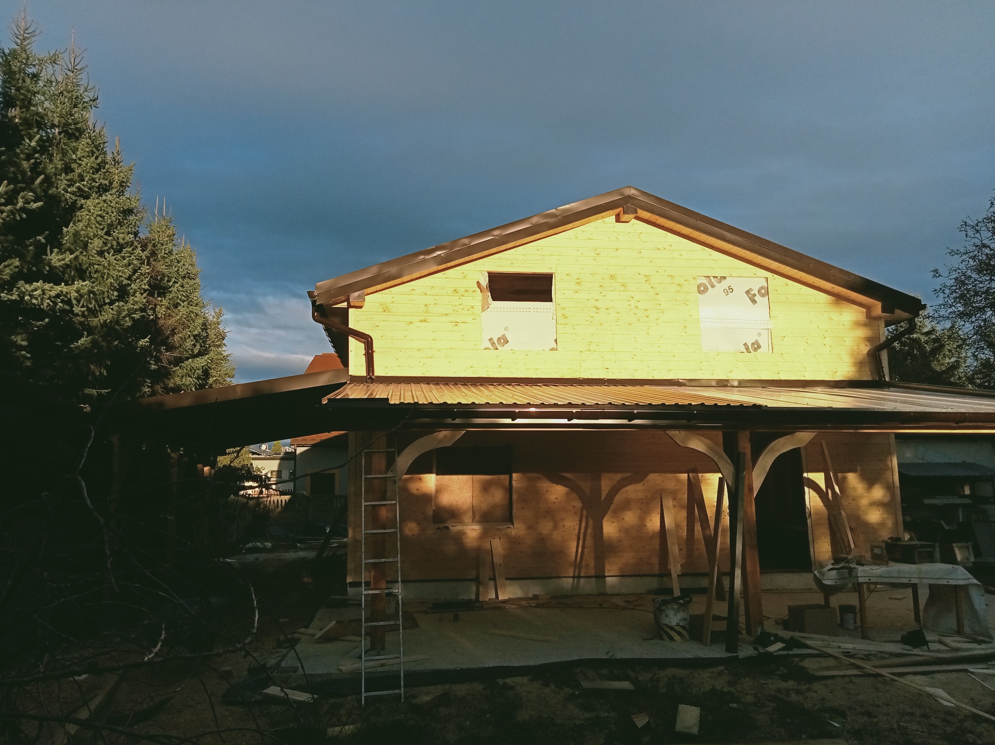 Dom w trakcie budowy z drewnianą konstrukcją dachu i ścian, widoczne zabezpieczenia okien, drabina oparta o ścianę, drzewa w tle i pochmurne niebo.