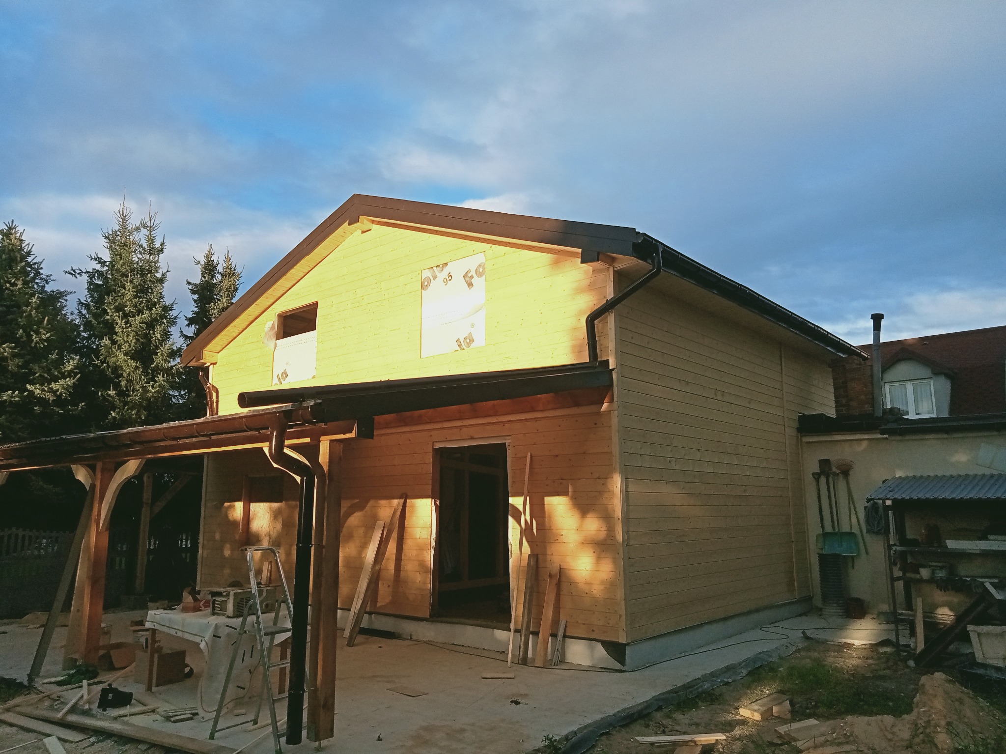 Dom w trakcie budowy z drewnianą elewacją i widocznymi elementami dachu, rynnami. Częściowo zakryte okna. Widoczne narzędzia i materiały budowlane.