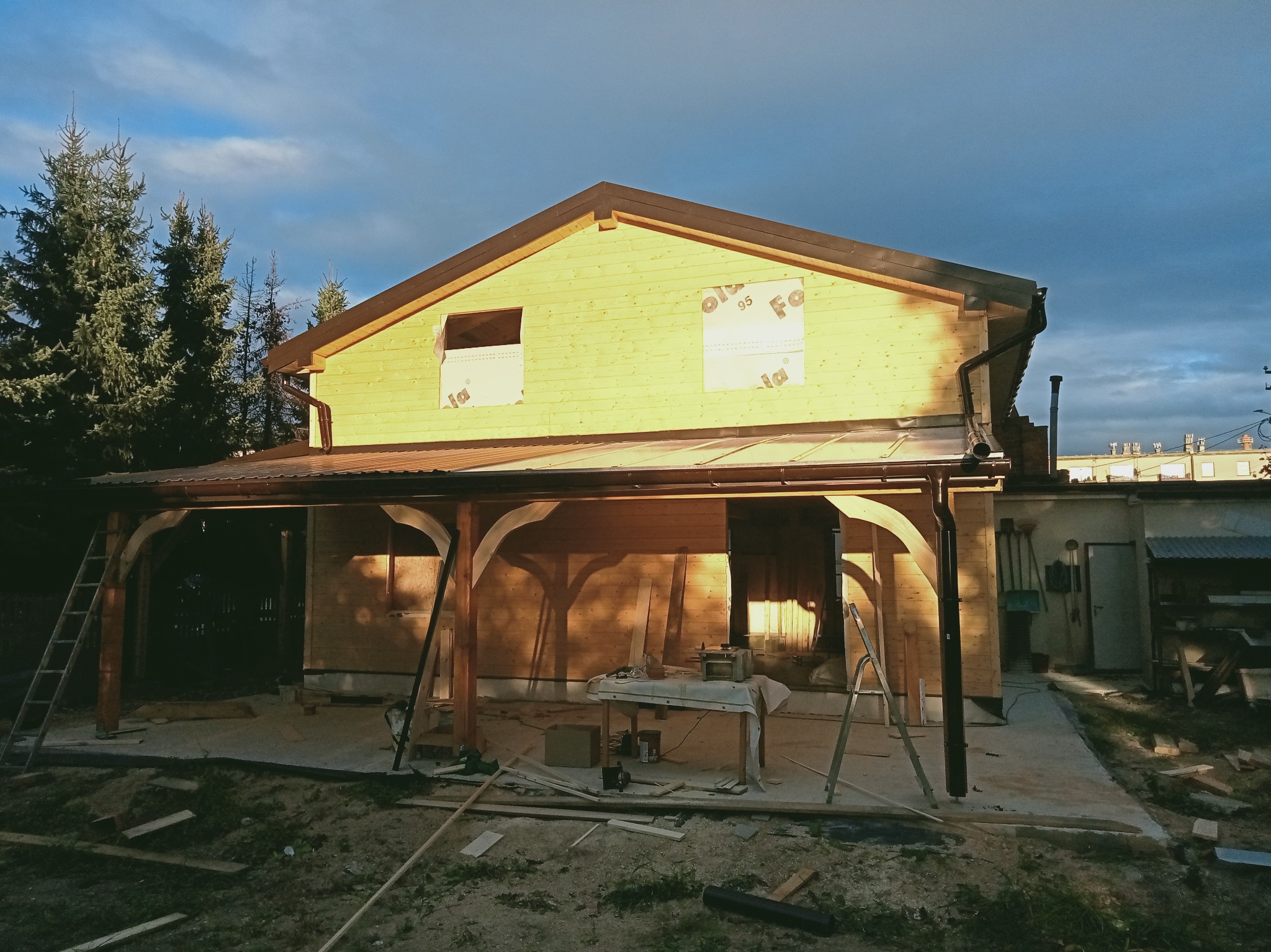 Żółty dom w trakcie budowy z widoczną więźbą dachową, oknami zabezpieczonymi folią, rynnami i elementami drewnianymi. Na pierwszym planie narzędzia i resztki materiałów budowlanych.