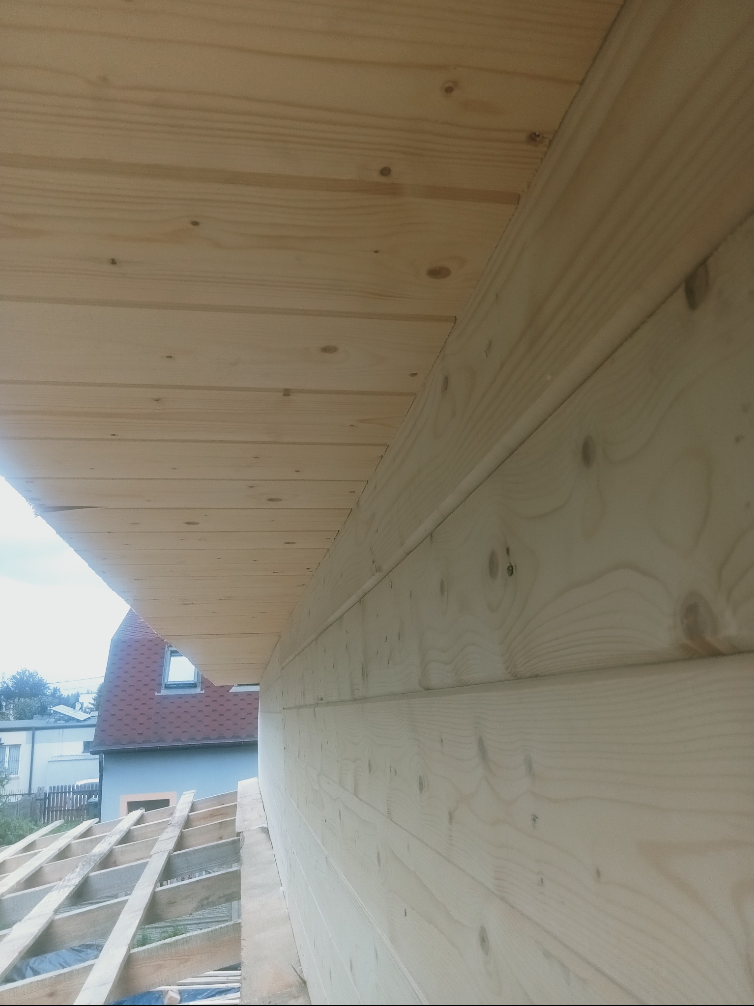 Drewniana podbitka dachowa z widoczną konstrukcją dachu w Opatowie. Jasne drewno, świeżo po montażu, zarys budynku w tle. Ujęcie z dołu, podkreślające detale.