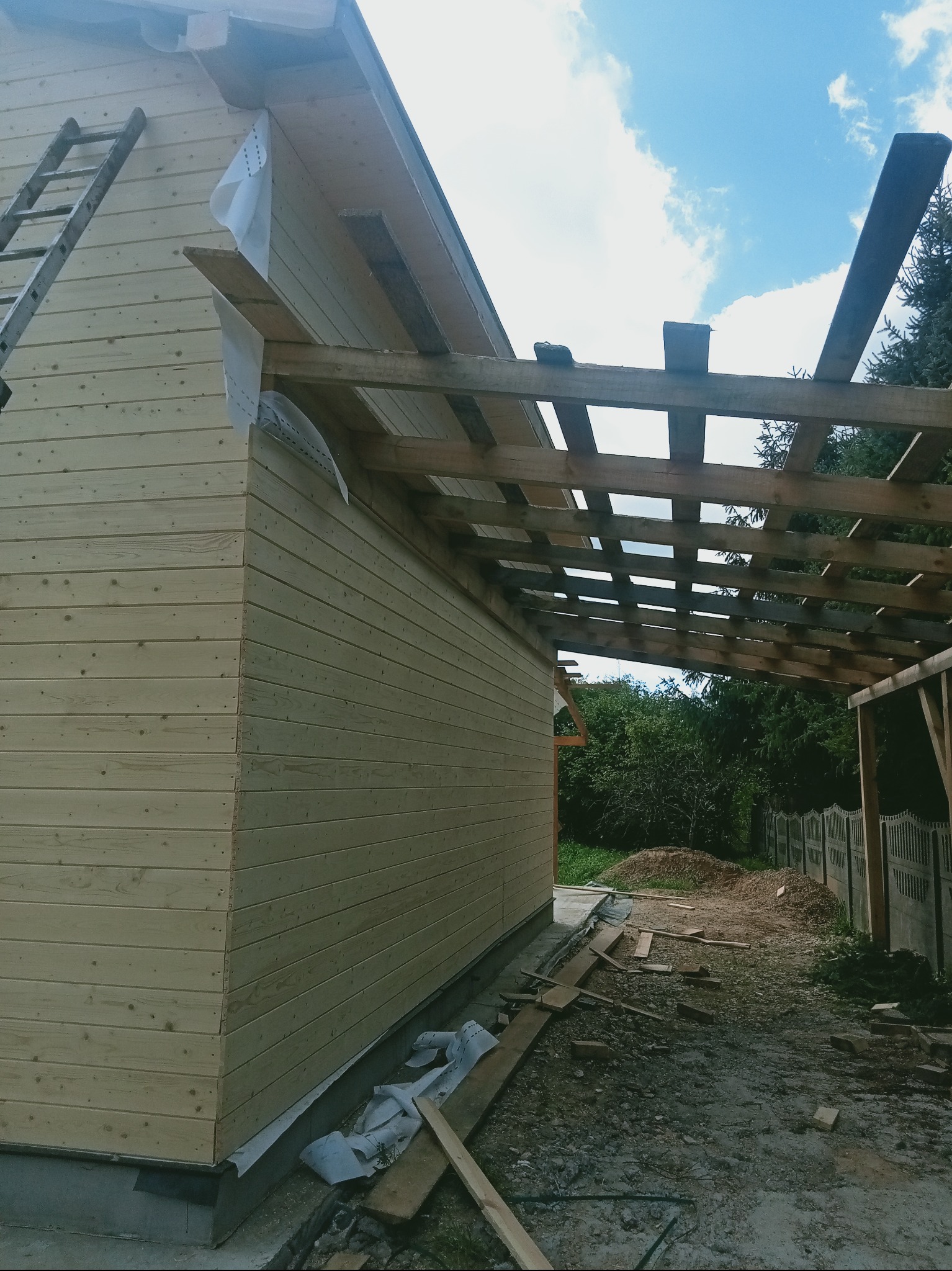Budowa drewnianej konstrukcji z widoczną więźbą dachową i deskowaniem ścian. Na ziemi leżą deski i inne materiały budowlane. W tle zieleń i błękitne niebo.