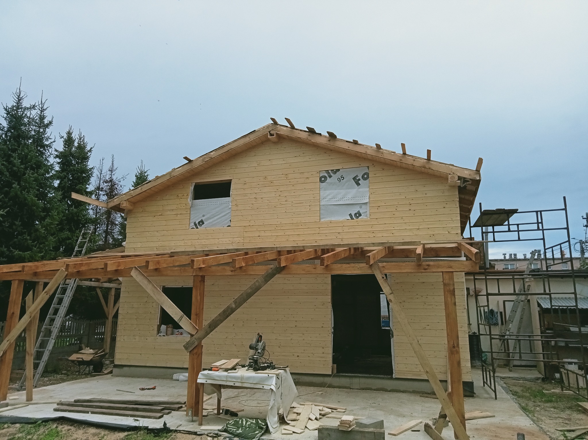 Surowy drewniany dom w trakcie budowy, widoczne elementy więźby dachowej, okna zabezpieczone folią, drabina oparta o ścianę, narzędzia na stole. Etap prac dekarskich.